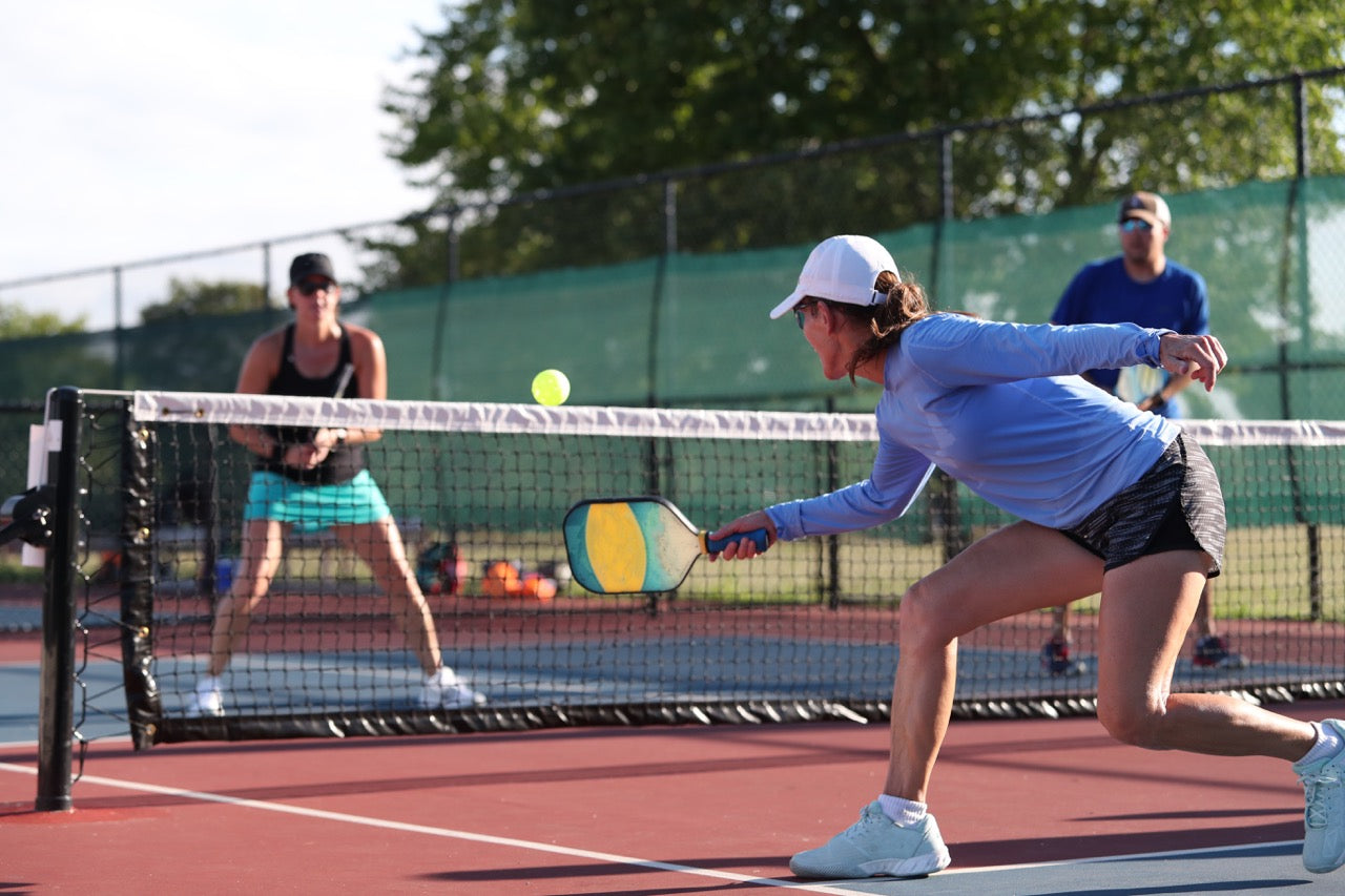 Group of people playing pickleball at an outdoor pickleball court with a woman hitting the pickleball closest to camera.