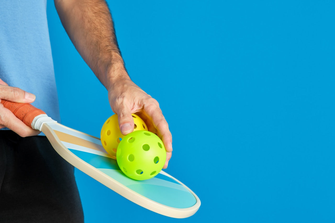 Man holding a pickleball paddle and two balls on a blue backdrop