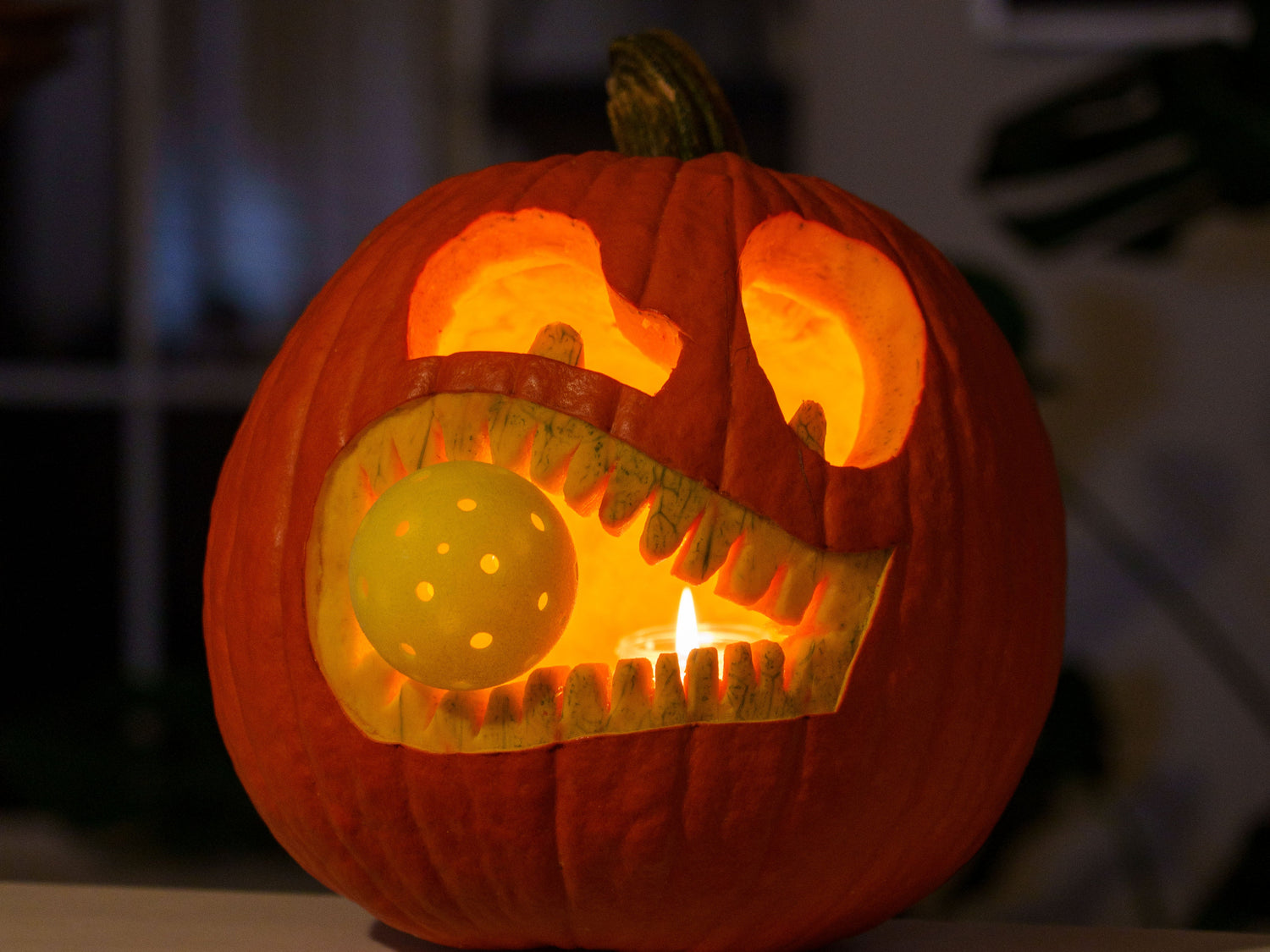 A pickleball themed jack o lantern carved out of a pumpkin featuring an animated face holding a pickleball in its mouth.