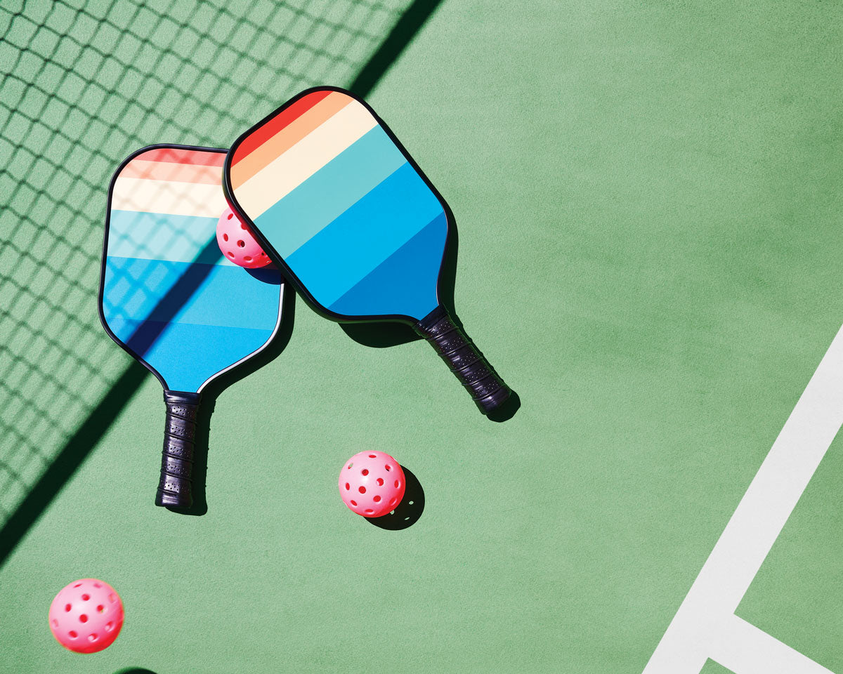 Two pickleball paddles and two pink balls on a pickleball court covered in the shadow of the net