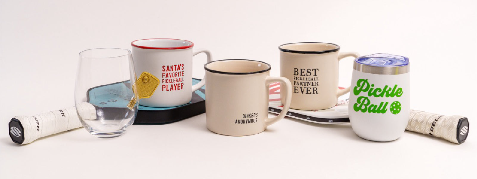 Five pickleball themed mugs and glasses sitting on two pickleball paddles in a studio.