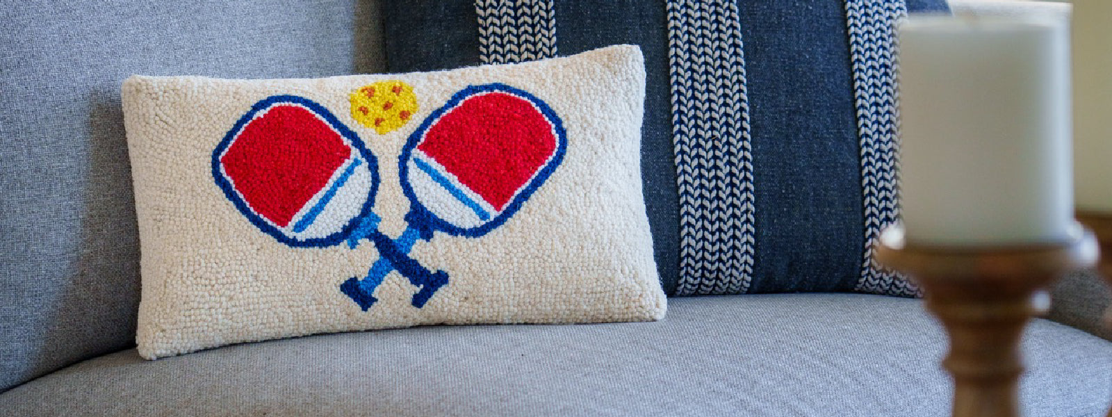 Cute hook pillow with two pickleball paddles and a pickleball placed on a couch with a candle in the foreground.