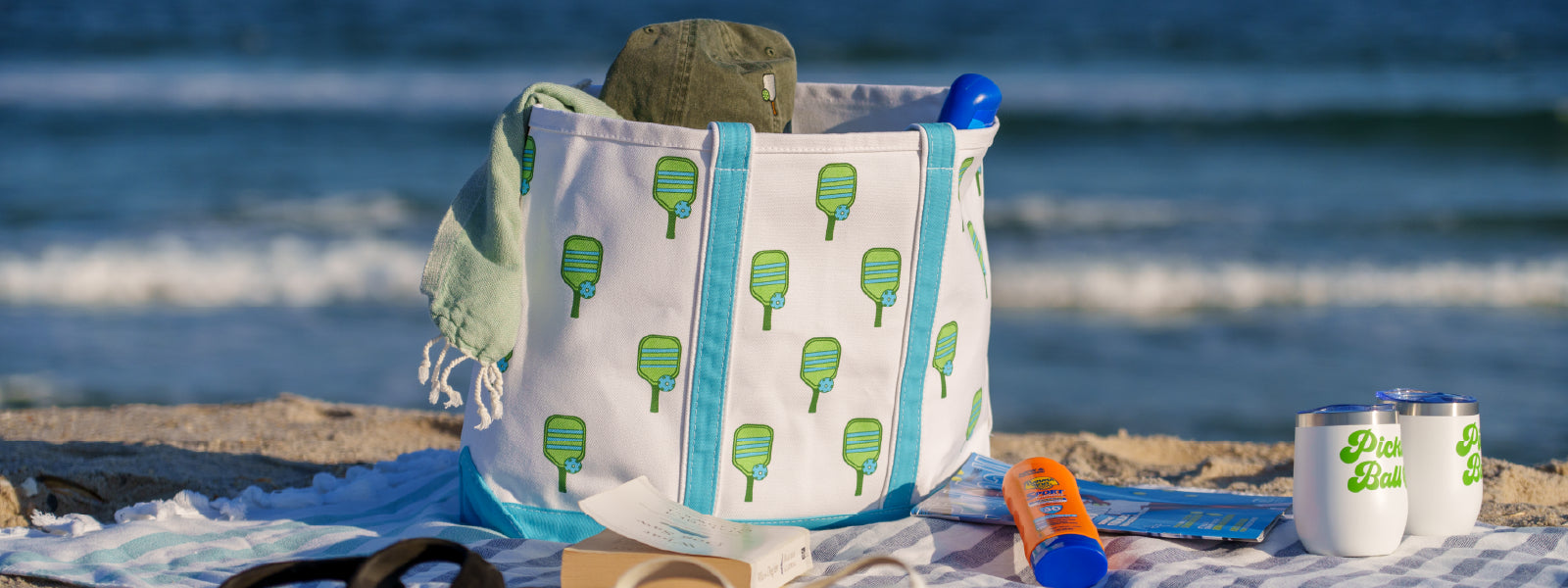 A tote bag with pickleball paddles on it sits on a beach towel in front of an ocean