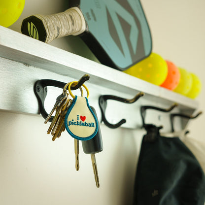 Keychain that says I Love Pickleball hanging on a coat hook with a pickleball paddle and balls in the background