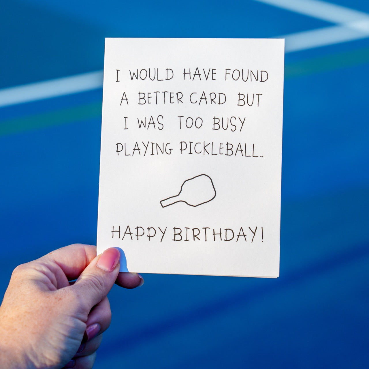 Person holding a funny pickleball birthday card that says &quot;I would have found a better card but I was too busy playing pickleball... Happy Birthday&quot; with a crudely drawn pickleball paddle in front of a pickleball court