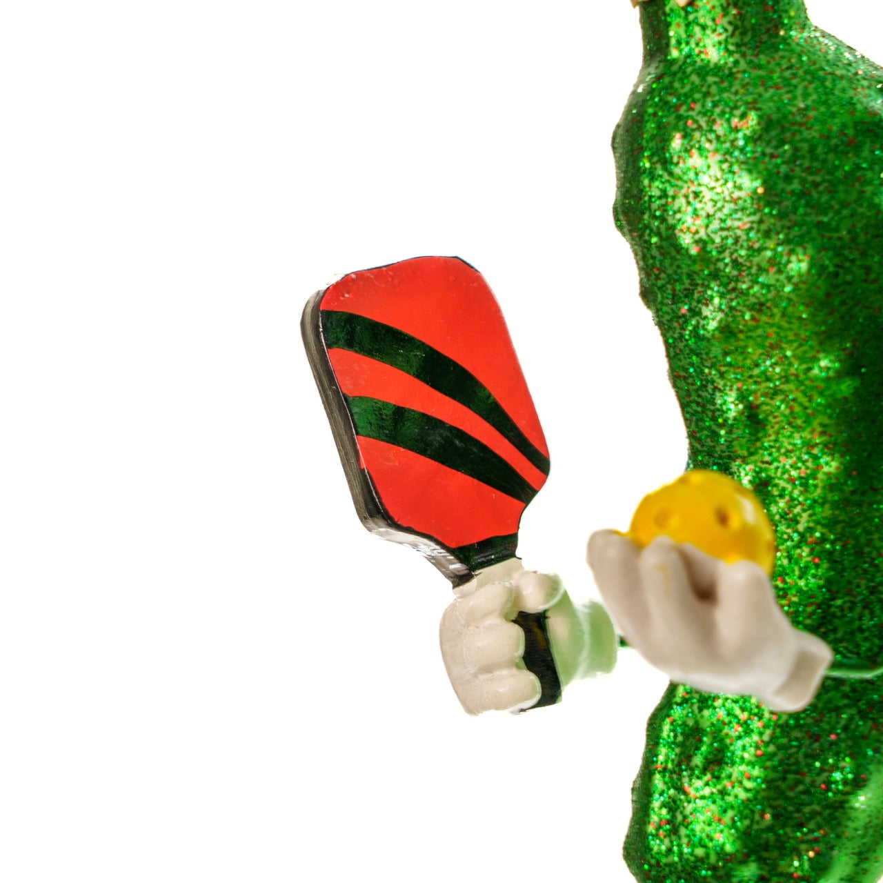 Close up of a pickleball paddle on a handmade Kurt Adler Christmas ornament of a pickle playing pickleball on a white studio backdrop