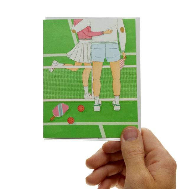 Pickleball card with the image of a couple hugging on a pickleball court being held.