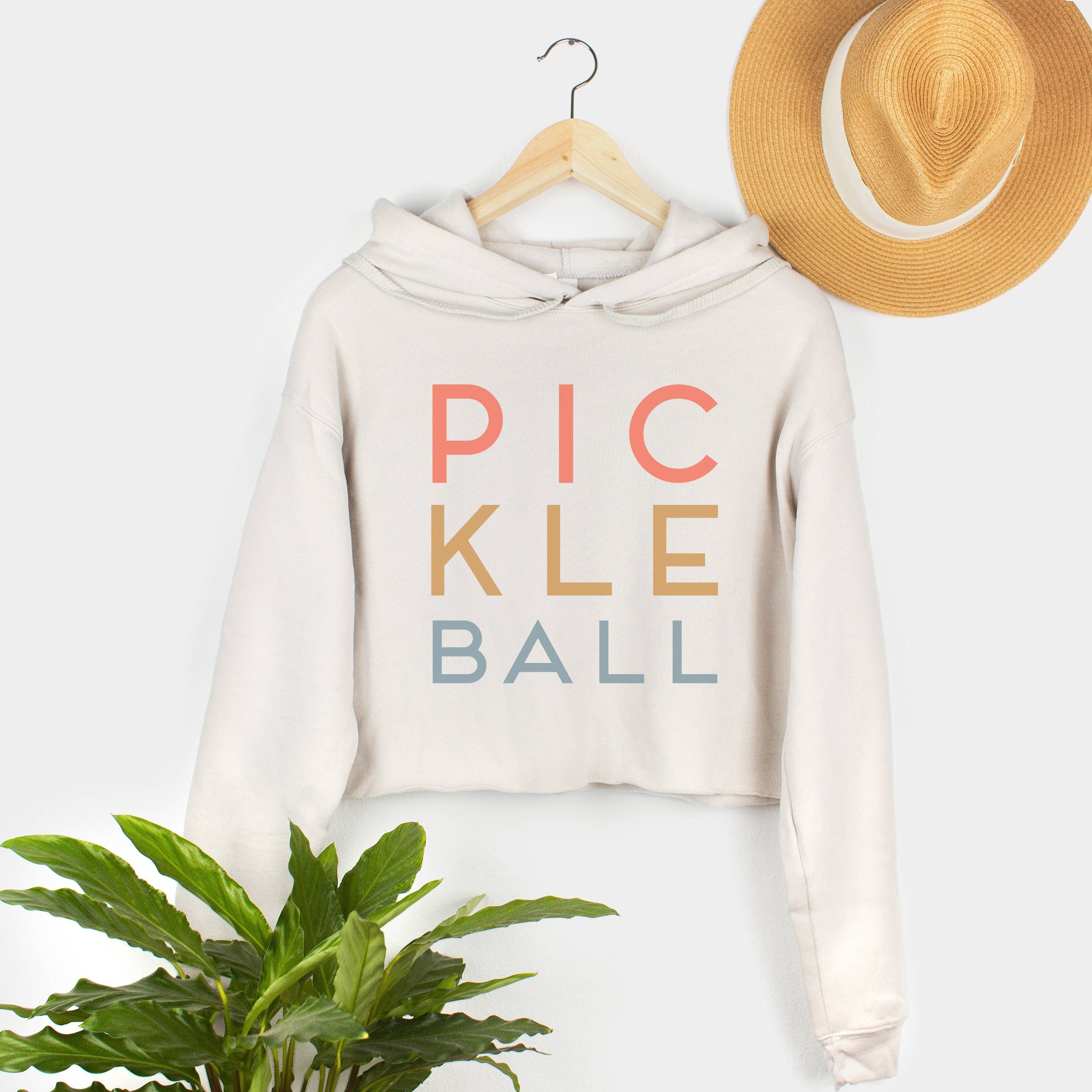 Hanging cream colored cropped hoodie sweatshirt that says &quot;PICKLEBALL&quot; on it in colorful lettering with a straw hat hanging above it and a plant sitting below it