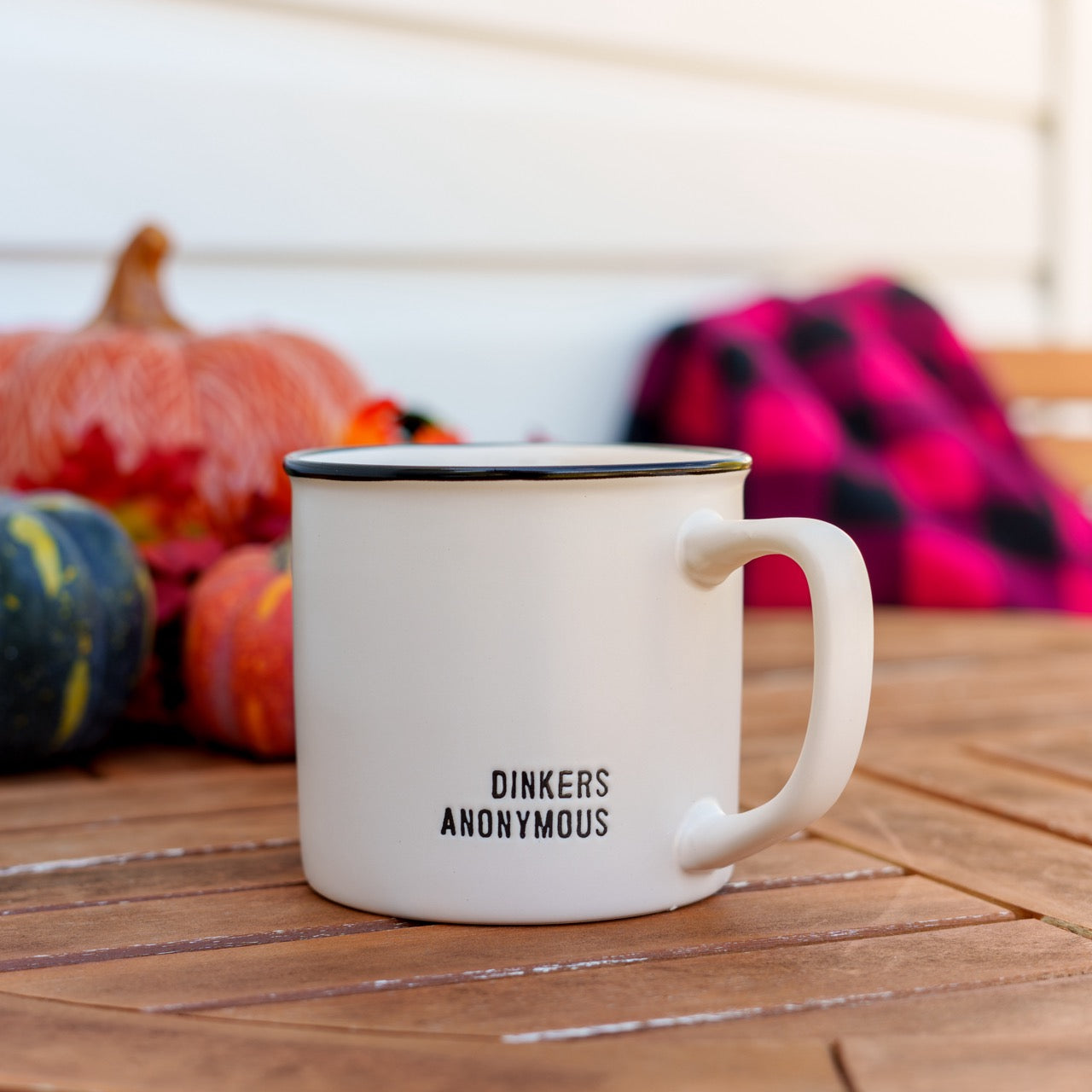 Stoneware coffee mug that says Dinkers Anonymous on a table in front of pumpkins and a blanket