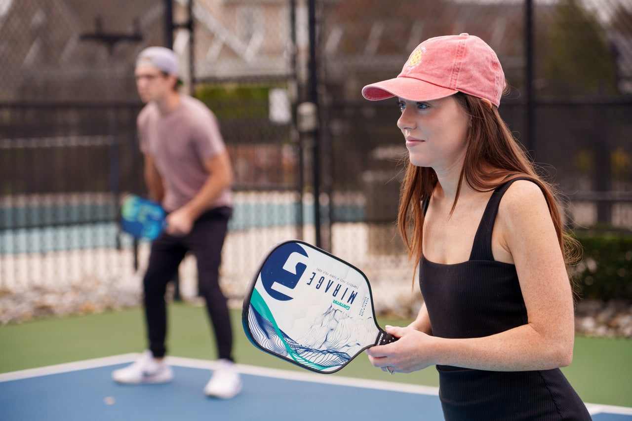 A man and a woman playing doubles pickleball with the woman wearing a red pickleball-themed baseball hat