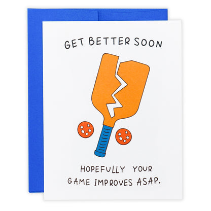 Funny pickleball themed get well soon greeting card that shows a broken paddle and two pickleballs with text that reads &quot;Get Better Soon, Hopefully your game improves ASAP.&quot; laying flat on a studio backdrop.