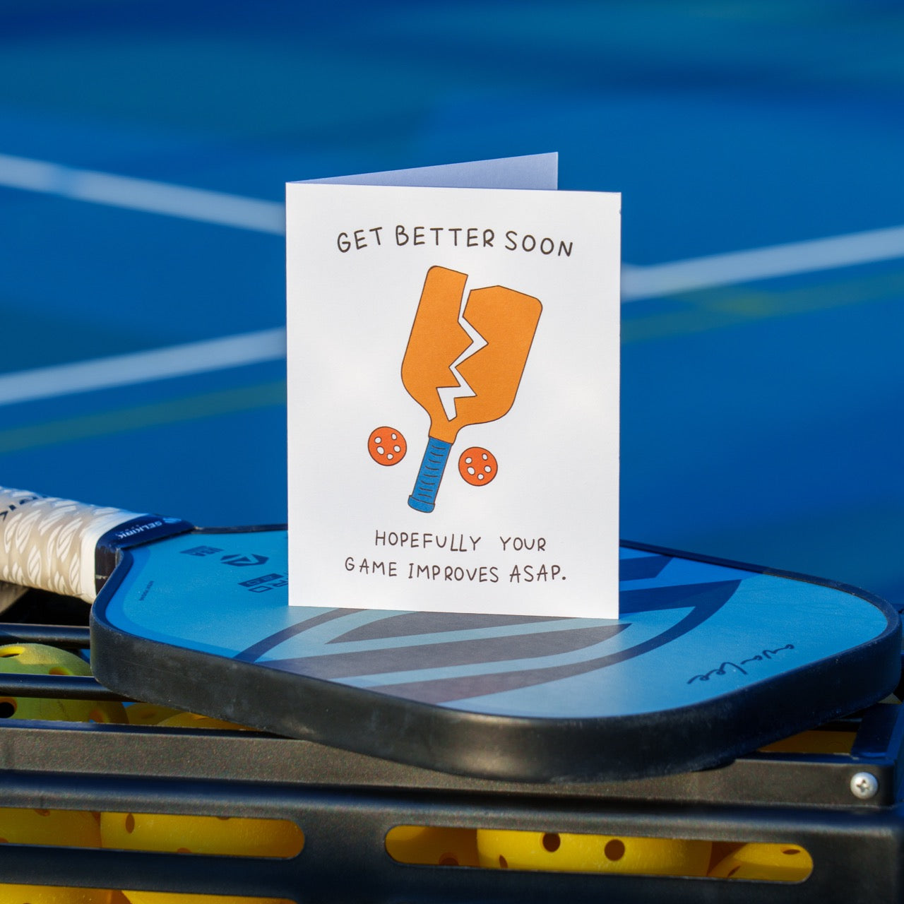 A funny pickleball themed get well soon greeting card that shows a broken paddle and two pickleballs with text that reads &quot;Get Better Soon, Hopefully your game improves ASAP.&quot; standing up on a pickleball paddle on a pickleball court.