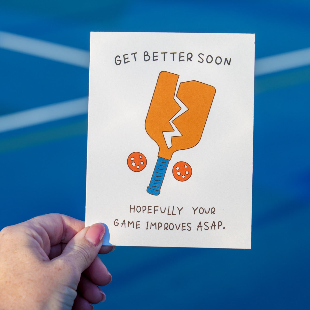 Person on a pickleball court holding a funny pickleball themed get well soon greeting card that shows a broken paddle and two pickleballs with text that reads &quot;Get Better Soon, Hopefully your game improves ASAP.&quot;