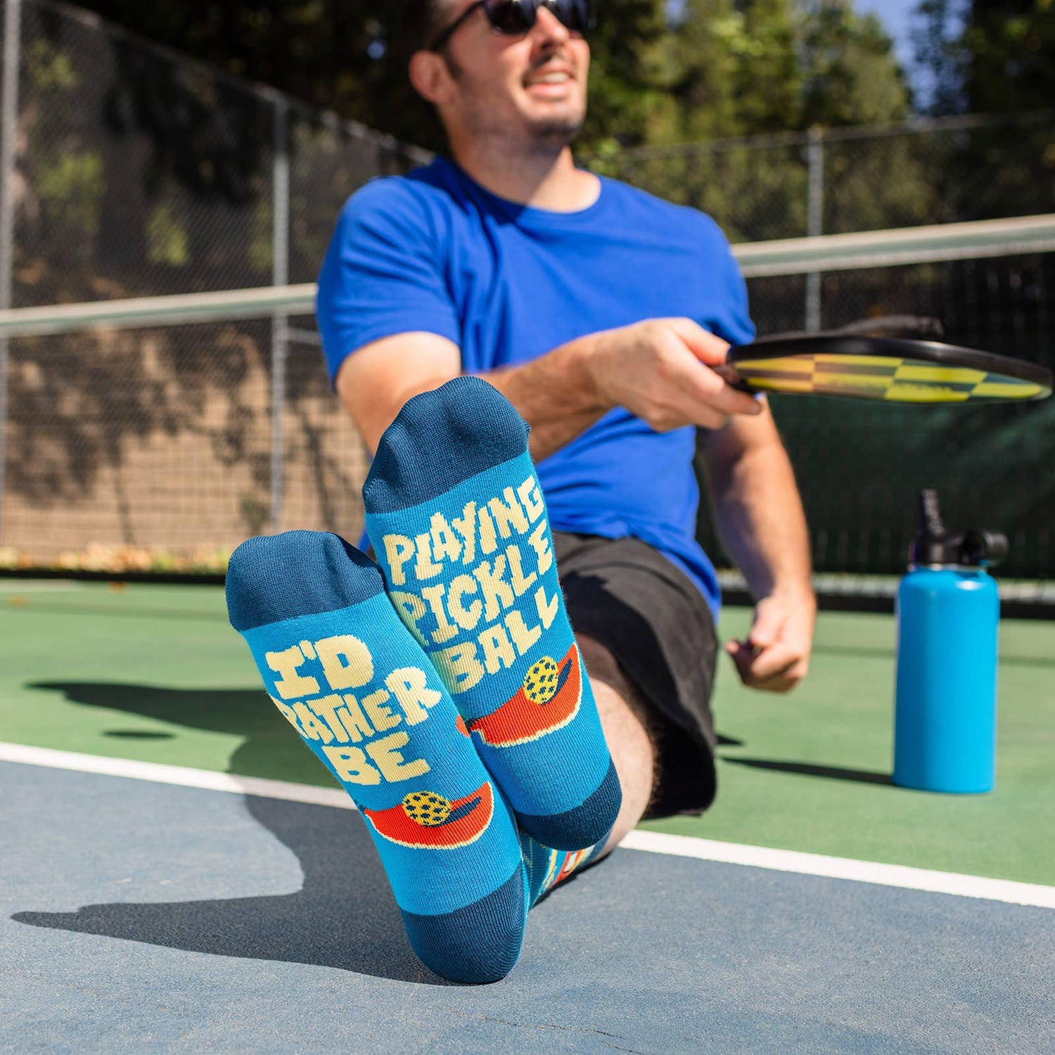 Man sitting on a pickleball court wearing humorous blue socks that say &quot;I&quot;D RATHER BE PLAYING PICKLEBALL&quot; on the soles of them