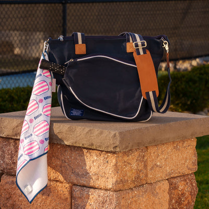 Navy pickleball tote bag with a pickleball themed sports towel hanging off of it sitting on a column in front of a pickleball court