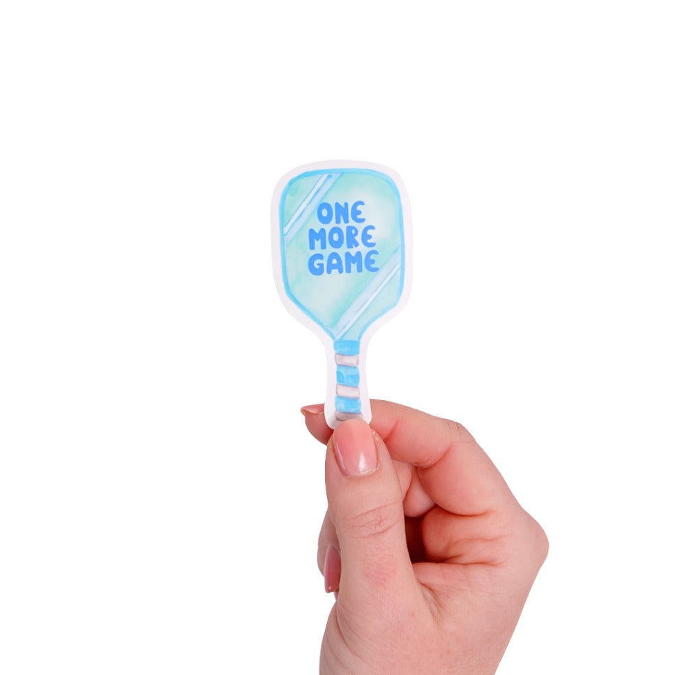 Blue sticker shaped like a pickleball paddle that says &quot;ONE MORE GAME&quot; on it being held