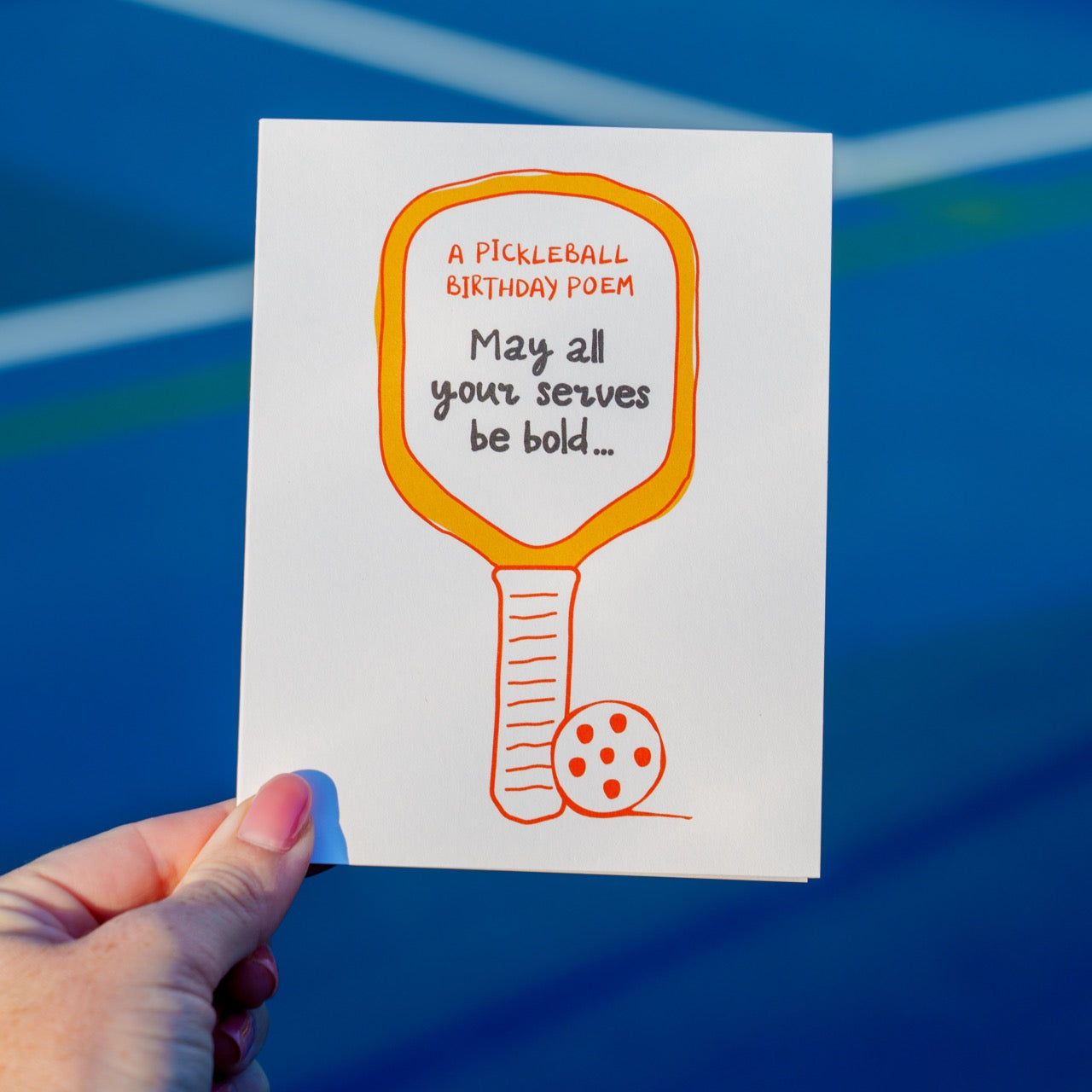 Person holding a funny pickleball birthday card with a pickleball paddle and ball that says &quot;A Pickleball Birthday Poem: May all your serves be bold...&quot; in front of a pickleball court