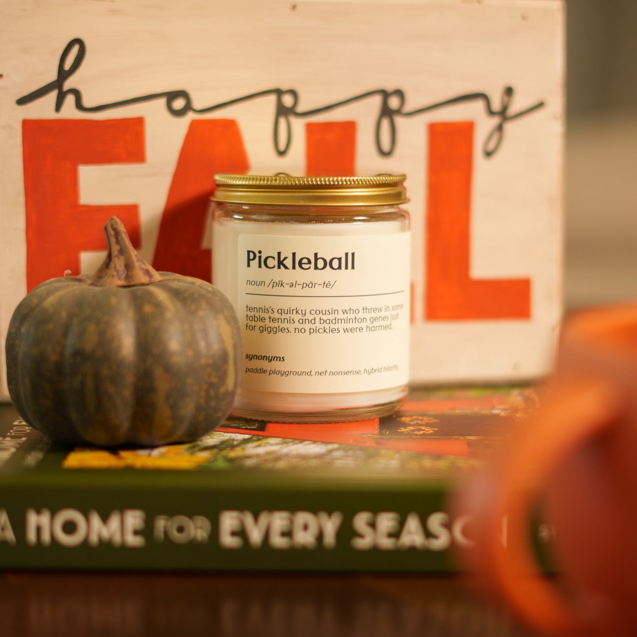 Pickleball definition candle sitting on a book with a pumpkin, coffee mug and a wooden sign that says FALL