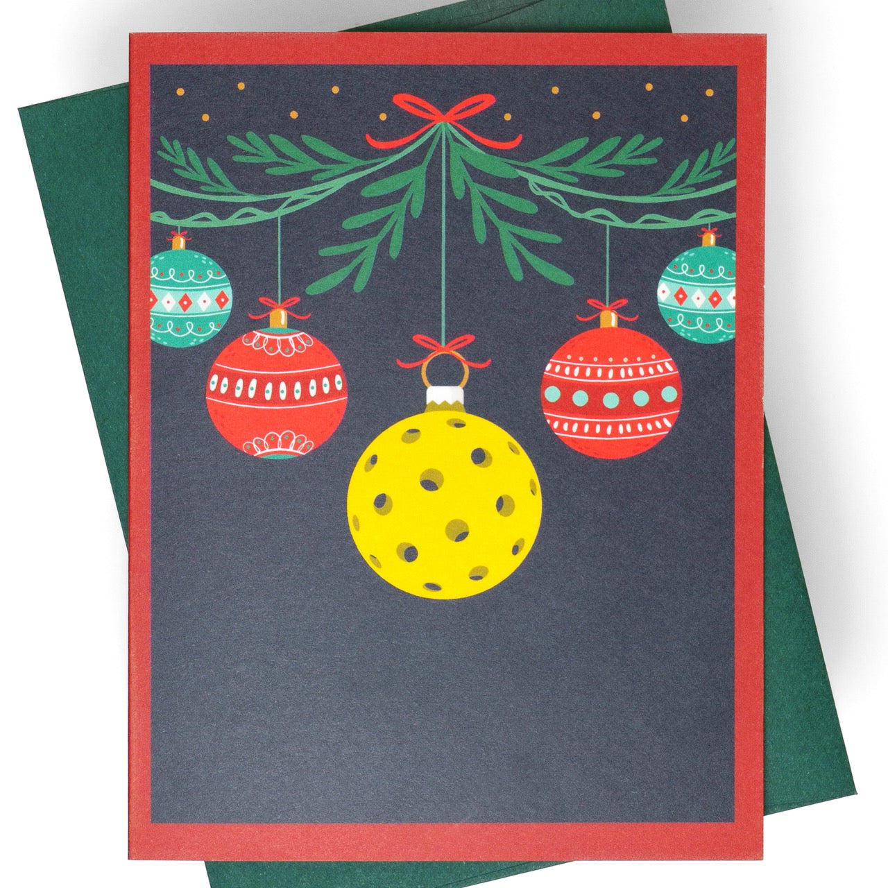 Close up of a pickleball themed Christmas greeting card with hanging ornaments and a pickleball with a green envelope laying flat