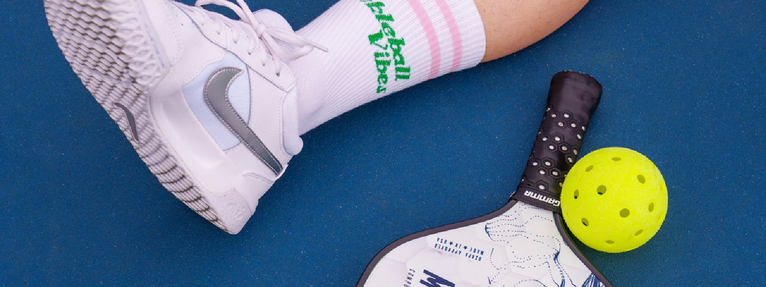 Woman sitting on the ground at a pickleball court with a close up on her paddle, pickleball, court shoes and trendy pickleball crew socks.