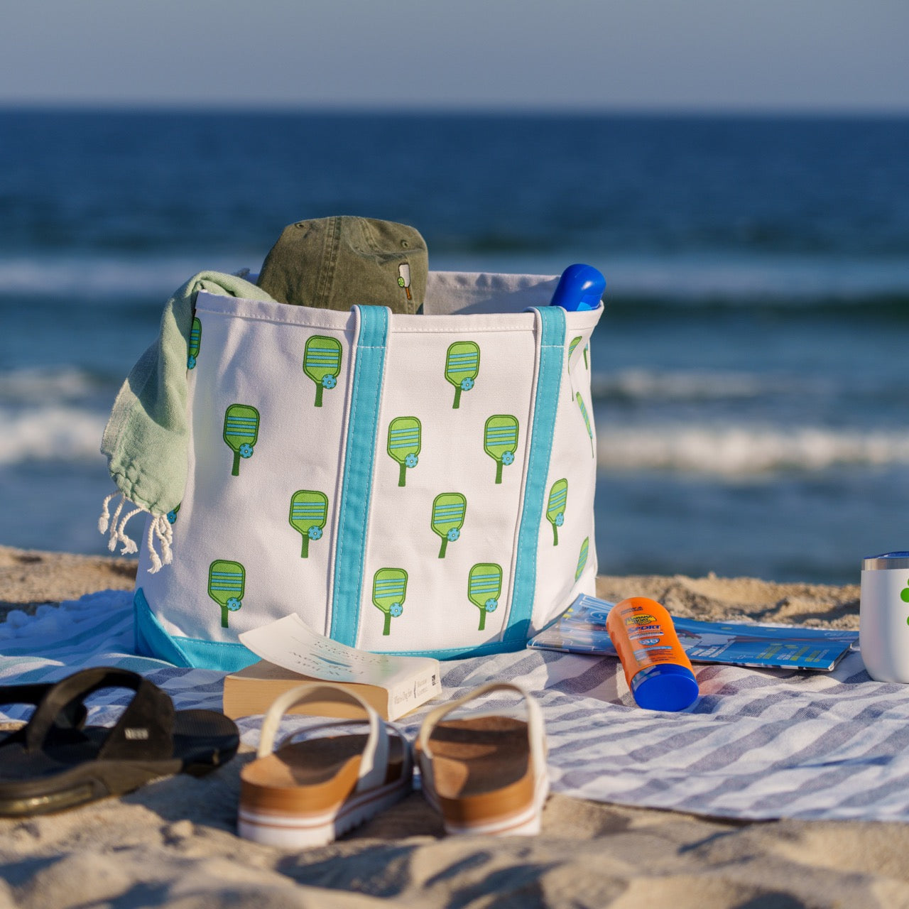 Collection of pickleball themed gifts, including a hat, tote bag and towel, on a beach