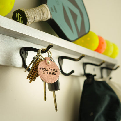 Pink leather keychain that says Pickleball Grandma hanging on a coat hook with a pickleball paddle and balls in the background