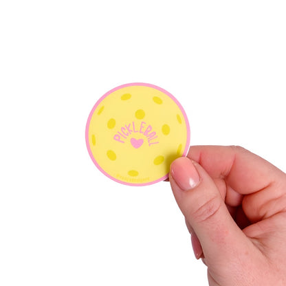 Yellow sticker shaped like a pickleball with a pink heart and pink &quot;PICKLEBALL&quot; on it being held