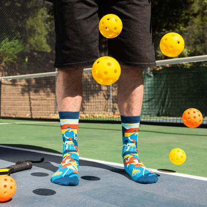 Person wearing humorous blue pickleball-themed socks on a pickleball court with pickleballs falling around them