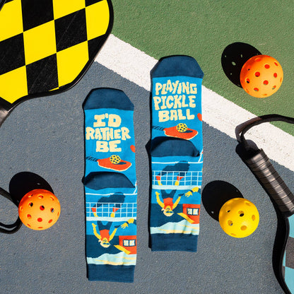 Blue humorous pickleball-themed socks on a pickleball court with balls and paddles surrounding them