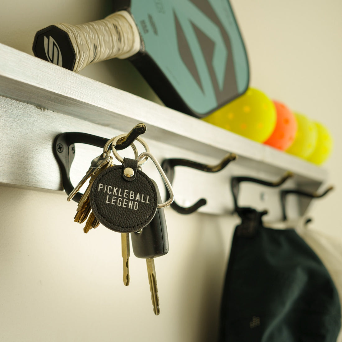 Black leather keychain that says Pickleball Legend hanging on a coat hook with a pickleball paddle and balls in the background