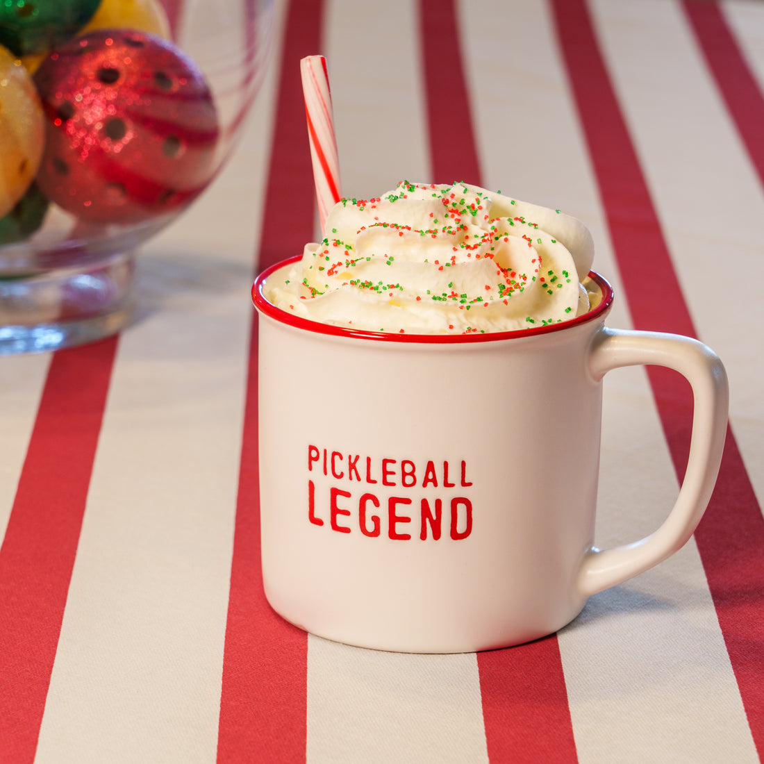 A white debossed mug that says &quot;PICKLEBALL LEGEND&quot; on it in red lettering with whipped cream and a candy cane in it sitting on a red and white striped table cloth