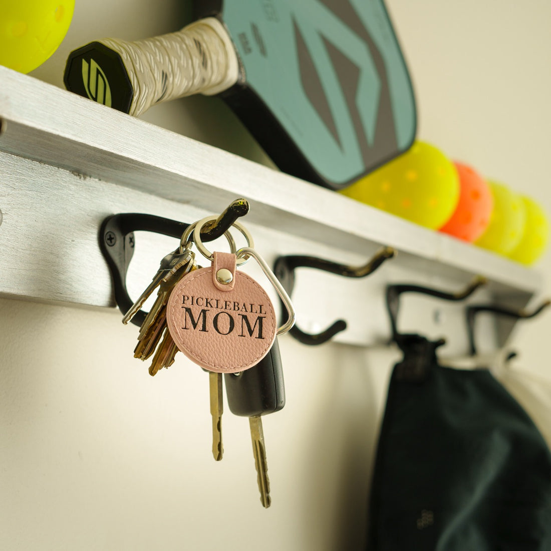 Pink leather keychain that says Pickleball Mom hanging on a coat hook with a pickleball paddle and balls in the background