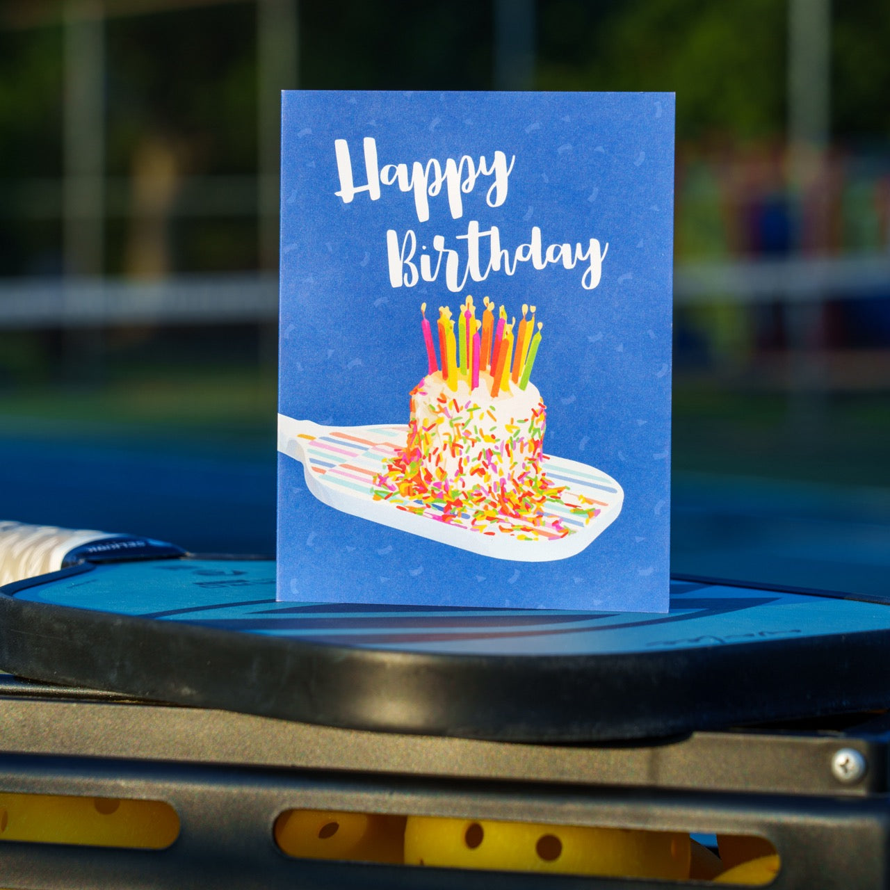 Colorful pickleball themed birthday card with a birthday cake with candles sitting on a pickleball paddle and big bold text that reads Happy Birthday standing on a pickleball paddle and pickleballs in front of a pickleball court.