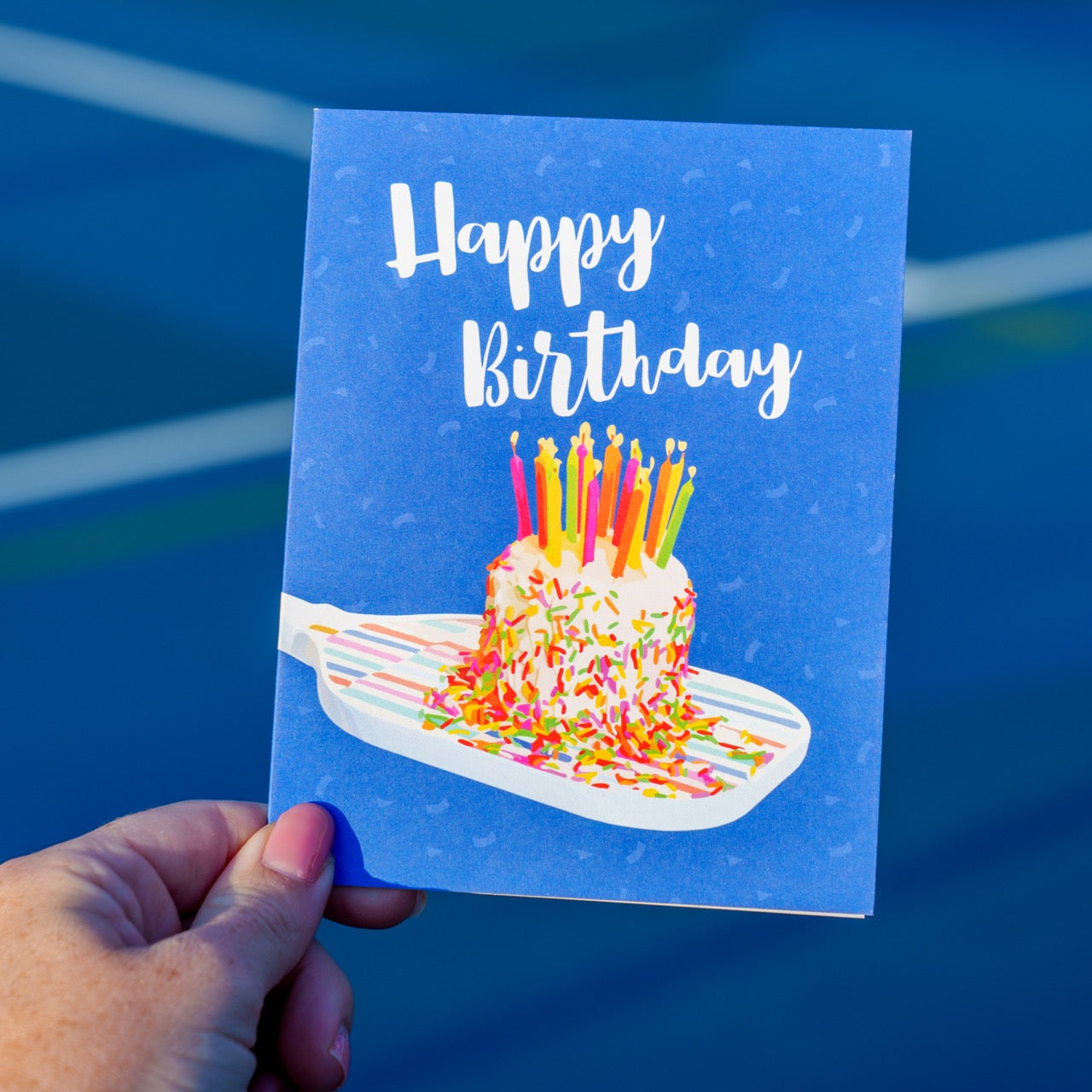 Person standing on a pickleball court holding a fun colorful pickleball themed birthday card with a birthday cake with candles sitting on a pickleball paddle and big bold text that reads Happy Birthday.