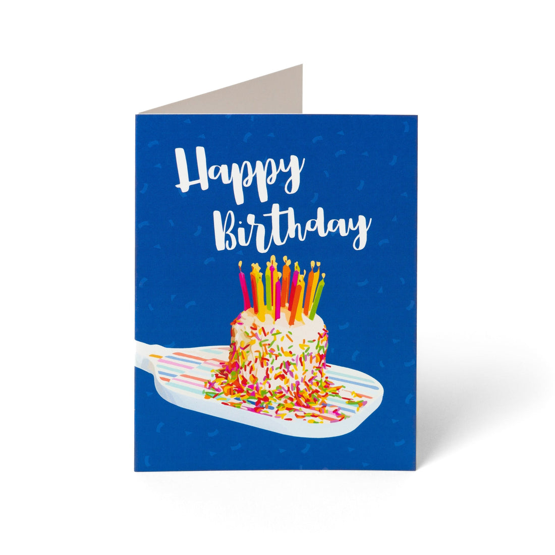 A fun colorful pickleball themed birthday card with a birthday cake with candles sitting on a pickleball paddle and big bold text that reads Happy Birthday standing up on a white background