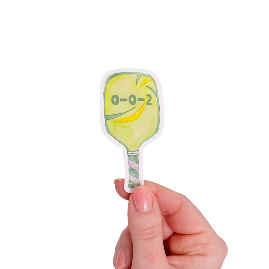 Green sticker shaped like a pickleball paddle with &quot;0-0-2&quot; on it being held