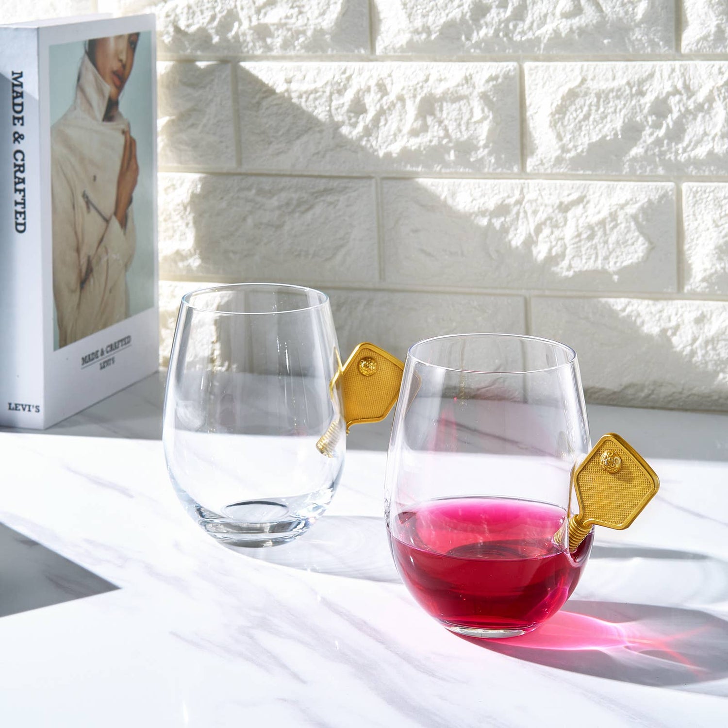 Set of stemless wine glasses with small metal pickleball paddles embedded into the glasses sitting on countertop with a book next to them