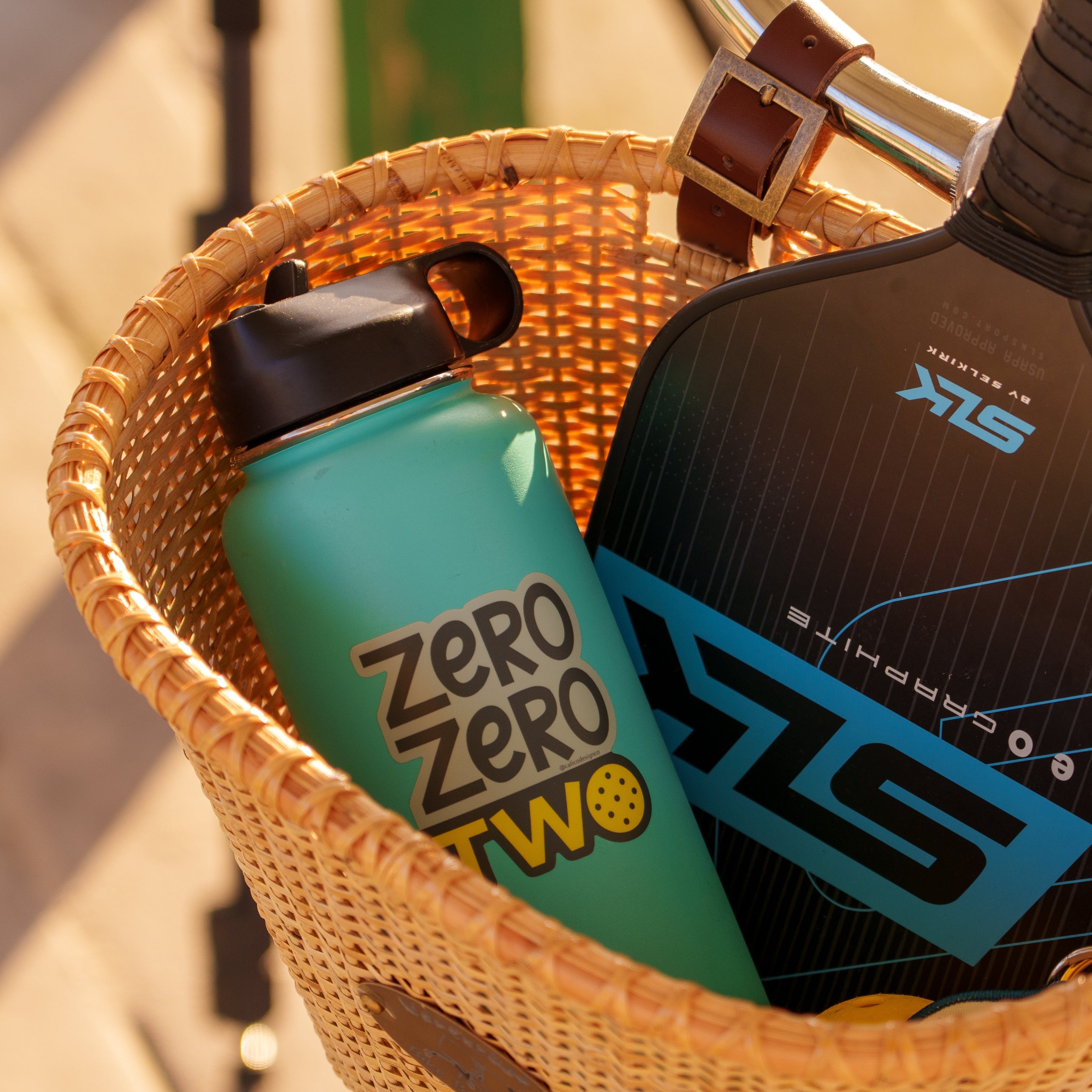 A water bottle and pickleball paddle in a bicycle basket with a sticker on a water bottle that says "ZERO ZERO TWO" on it