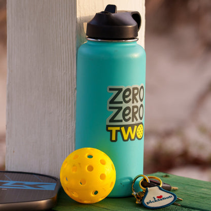 Blue water bottle with a pickleball-themed sticker on it that says &quot;ZERO ZERO TWO&quot; next to a pickleball paddle and ball on a boardwalk