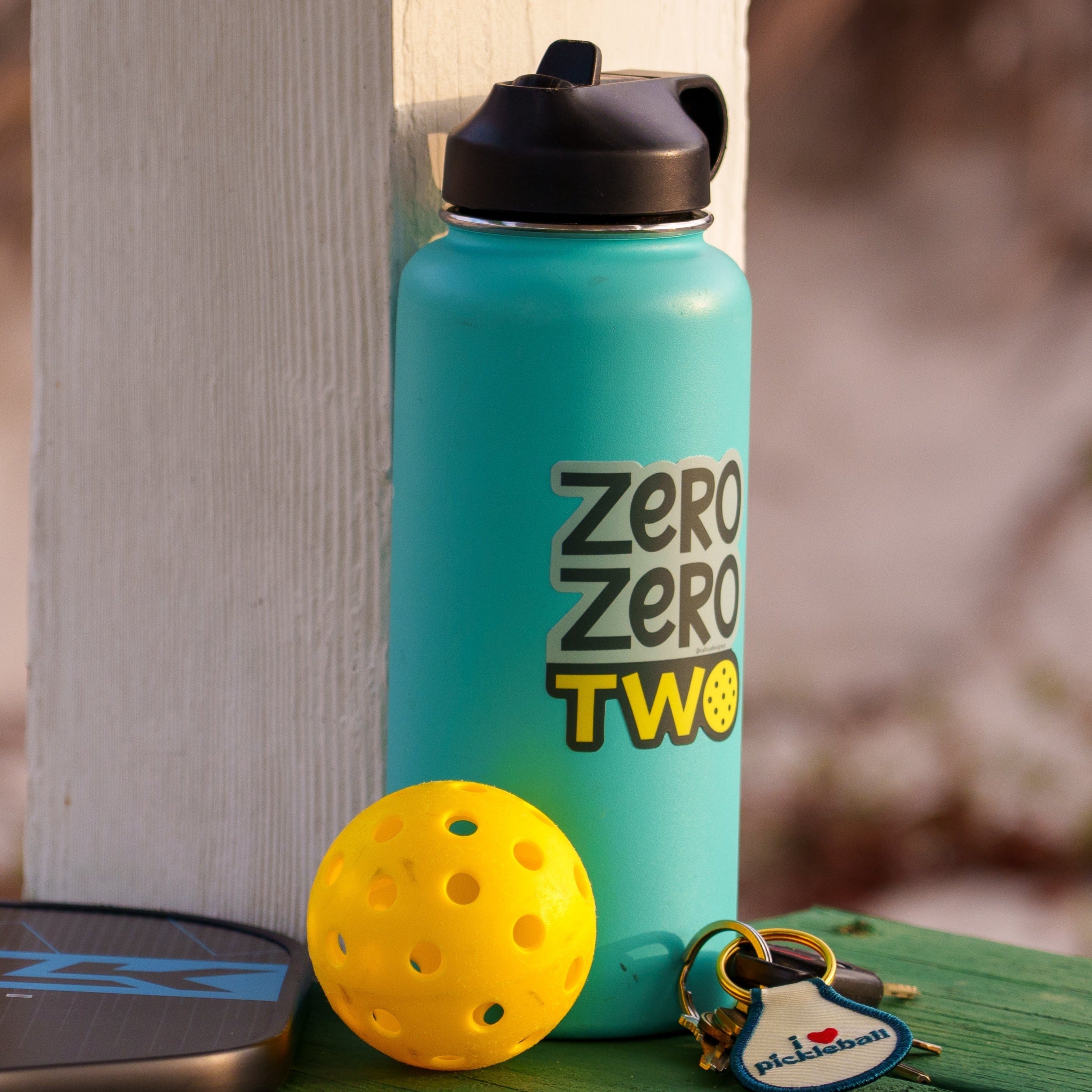 Blue water bottle with a pickleball-themed sticker on it that says &quot;ZERO ZERO TWO&quot; next to a pickleball paddle and ball on a boardwalk