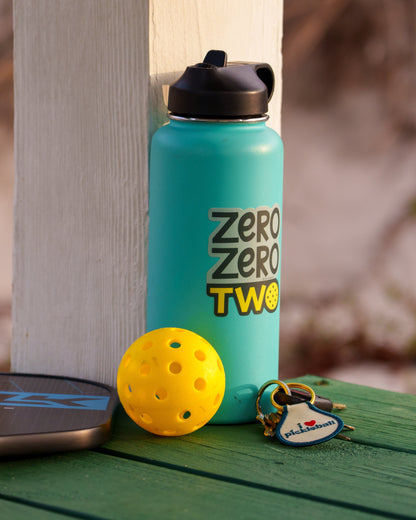 Blue water bottle with a pickleball-themed sticker on it that says &quot;ZERO ZERO TWO&quot; next to a pickleball paddle and ball on a boardwalk