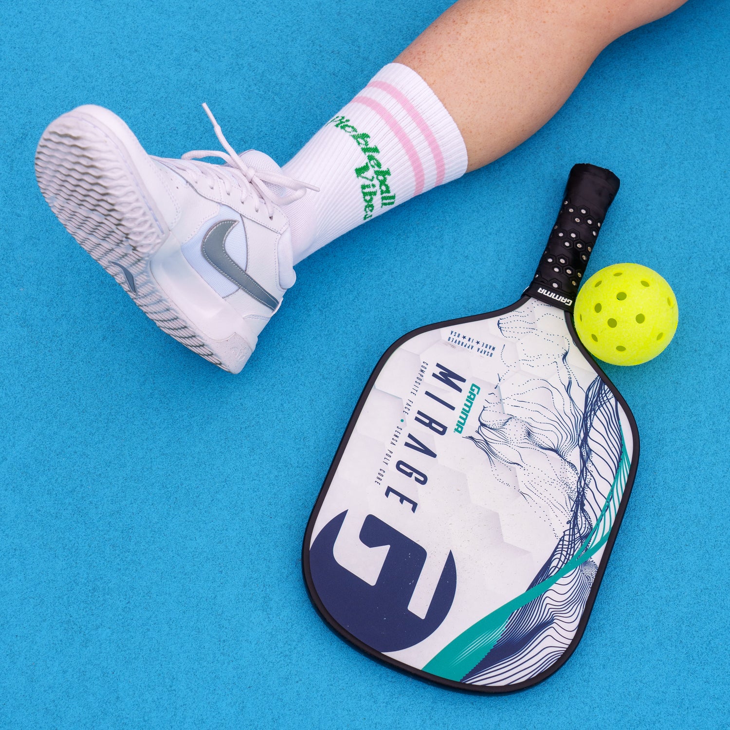 Person's leg on a light blue pickleball court wearing socks that say "Pickleball Vibes" in pink text with a paddle and ball next to their foot