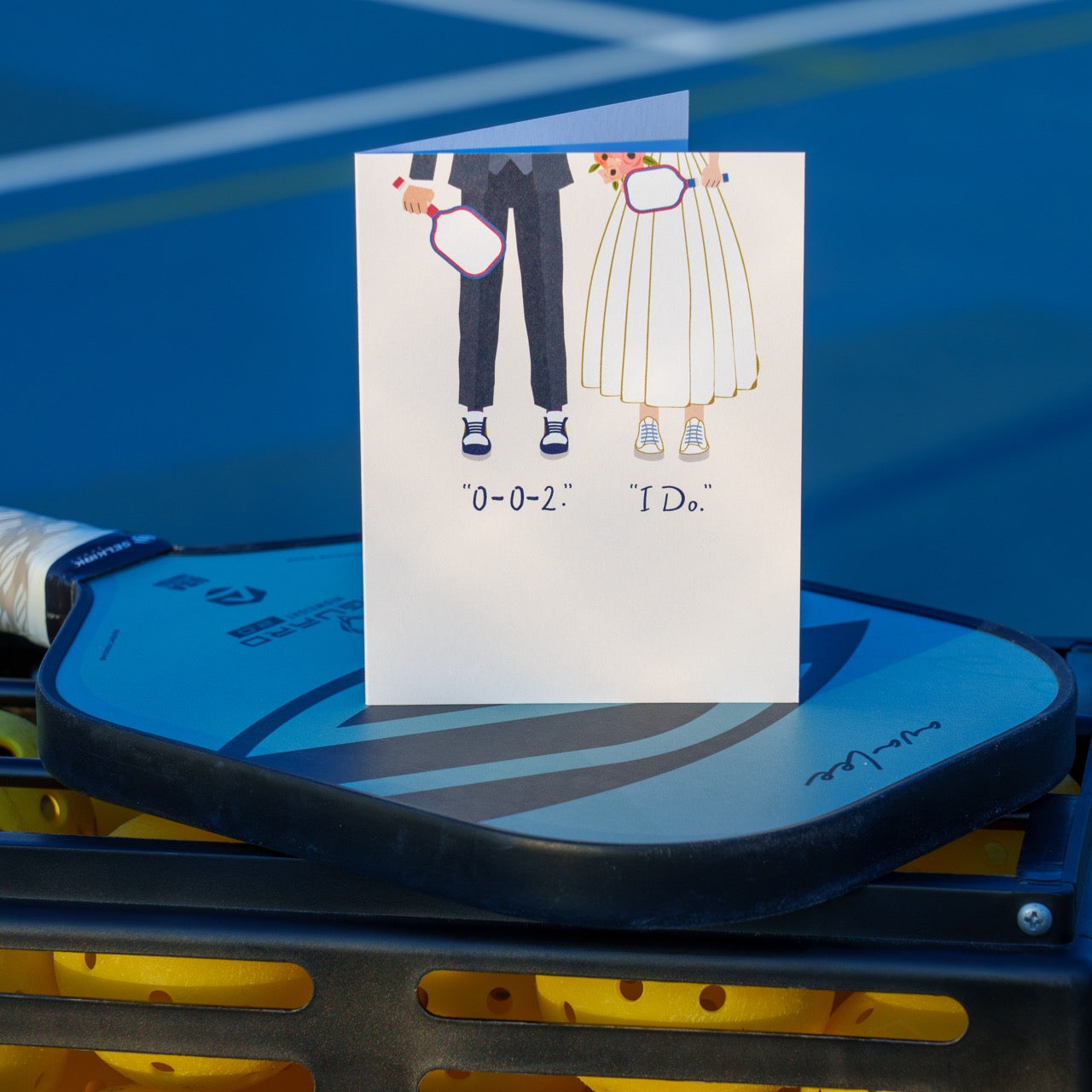 An elegant pickleball themed wedding card that shows a bride and a groom on their wedding day holding pickleball paddles wearing court shoes with text that reads &quot;0-0-2.&quot;, &quot;I Do.&quot;, standing up on a pickleball paddle at a pickleball court