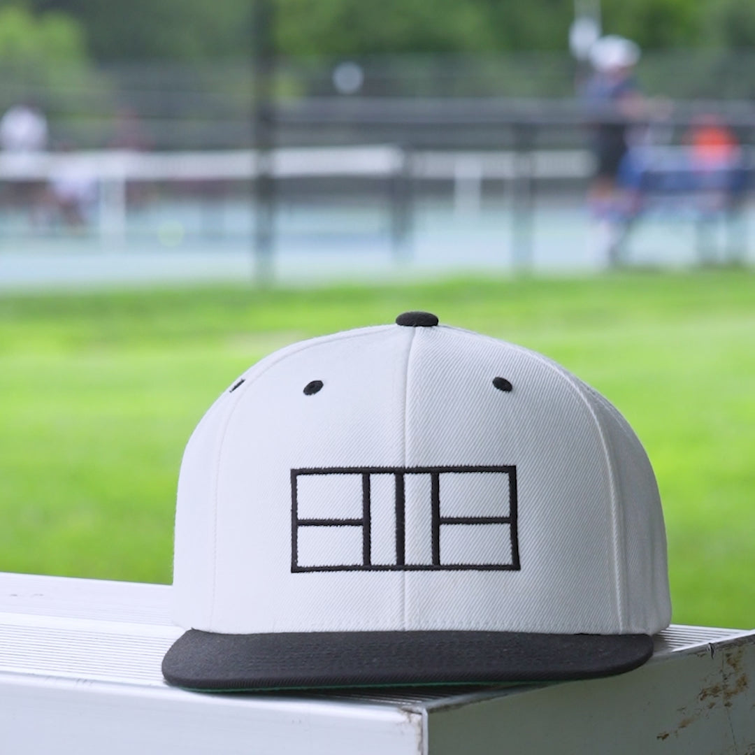 Video of a white and black snapback hat with a black embroidered pickleball court on it