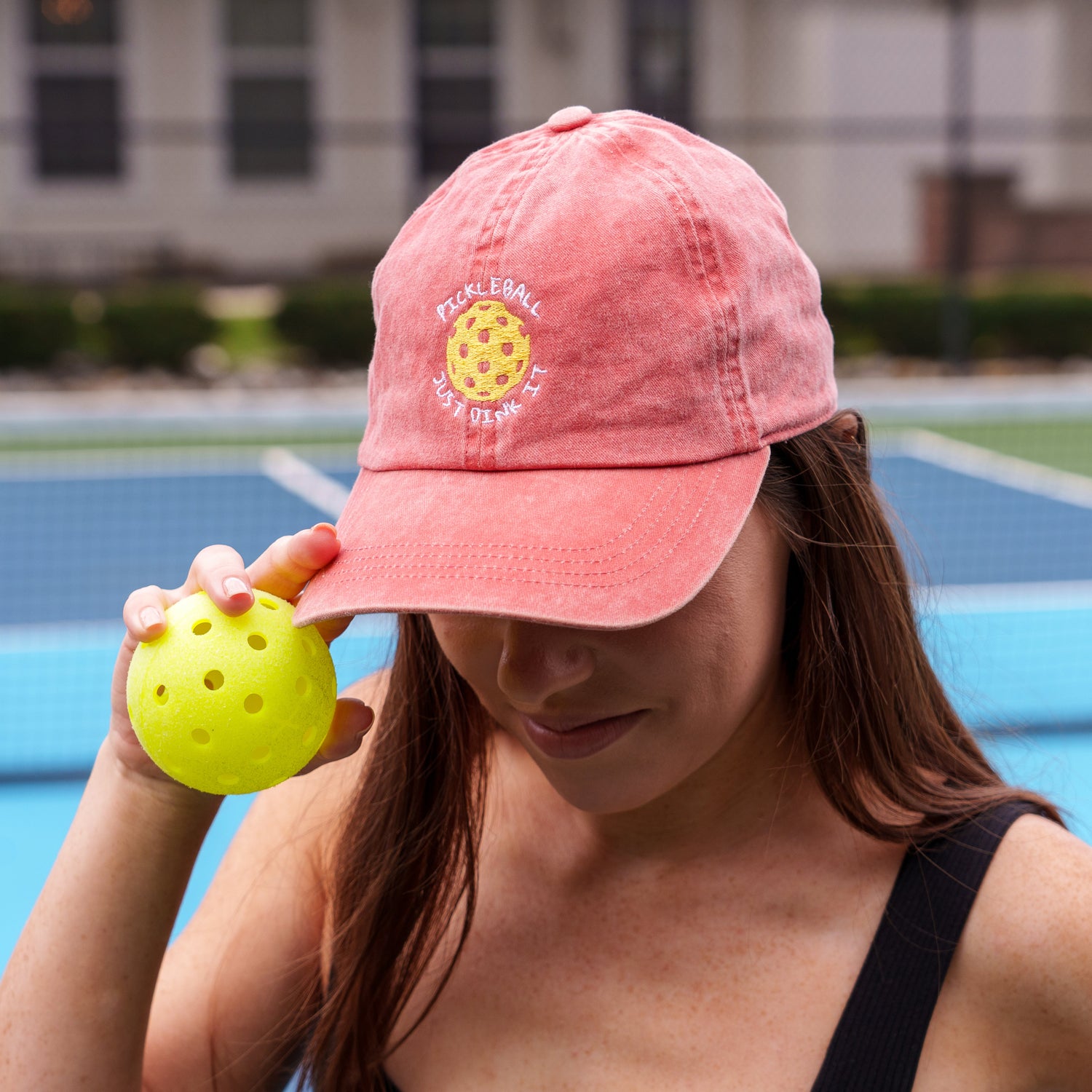 Woman on a pickleball court wearing a red embroidered hat that says &quot;PICKLEBALL JUST DINK IT&quot; on it and holding a pickleball