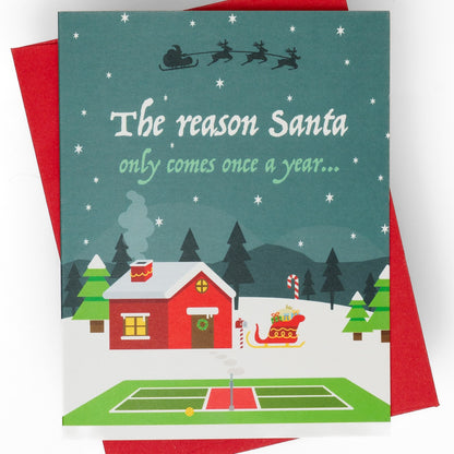 Close up front of a pickleball themed Christmas greeting card showing Santas home in the north pole with a pickleball court in the front with a message that says &quot;The reason Santa only comes once a year... is actually because he&