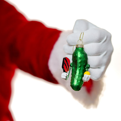 Santa holding a green glass pickle Christmas ornament that holding a pickleball paddle and ball in front of a white studio backdrop