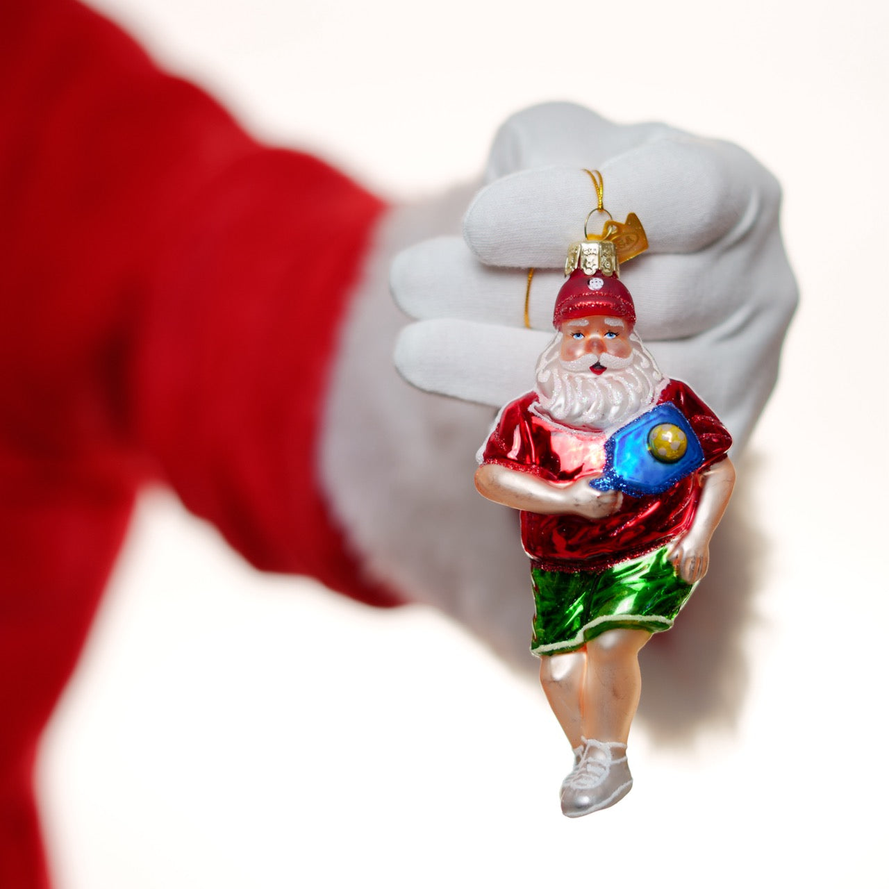 Santa holding a premium glass Christmas ornament of Santa with a pickleball paddle and ball standing in front of a white studio back drop