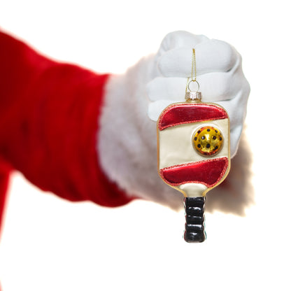 Santa holding a red glass Christmas ornament of a pickleball paddle and ball in front of a white studio backdrop