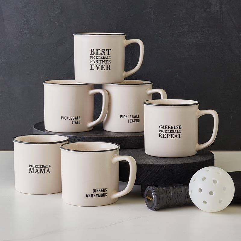 Collection of stoneware mugs with pickleball themed sayings, a white pickleball and pickleball paddle.